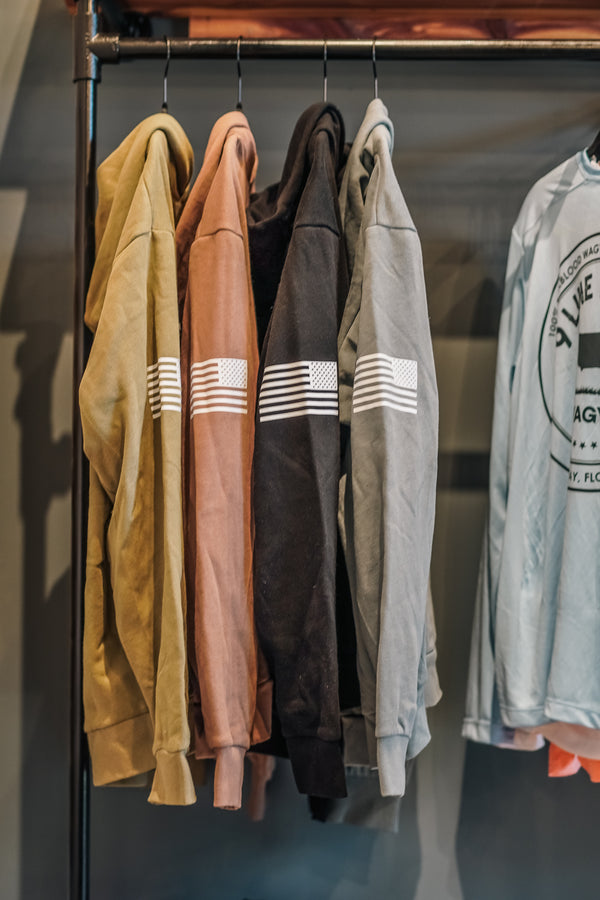 Four hooded sweatshirts in tan, blush pink, black, and heather gray hang on a black clothing rack. Each sweatshirt has a white horizontal stripe graphic with three vertical stripes on the upper sleeve.