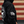A person wearing a black hoodie with a large white circular graphic on the back and a white striped flag-style patch on the sleeve, shown against a red and beige wood plank background.
