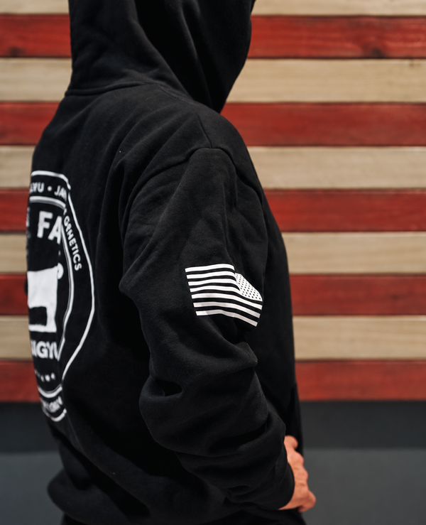 A person wearing a black hoodie with a large white circular graphic on the back and a white striped flag-style patch on the sleeve, shown against a red and beige wood plank background.