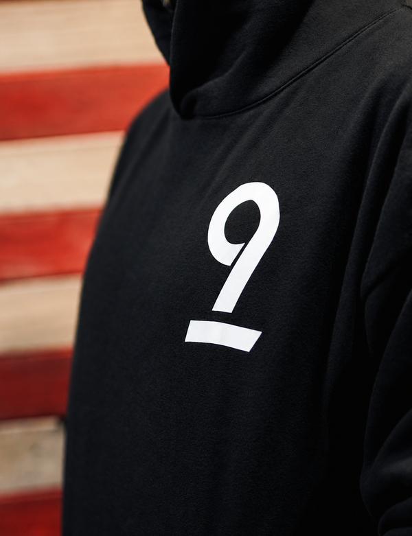 Close-up of a black hoodie with a large white number 9 printed on the chest, worn by a person in front of a blurred red and beige striped background.