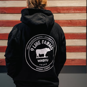 A person viewed from behind wearing a black hoodie with a large white circular graphic on the back that reads '9 Line Farms Wagyu' and shows a cow silhouette. The background is a rustic wooden wall with horizontal red and beige wood planks.