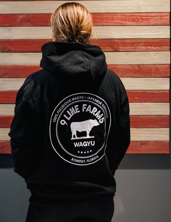 A person viewed from behind wearing a black hoodie with a large white circular graphic on the back that reads '9 Line Farms Wagyu' and shows a cow silhouette. The background is a rustic wooden wall with horizontal red and beige wood planks.