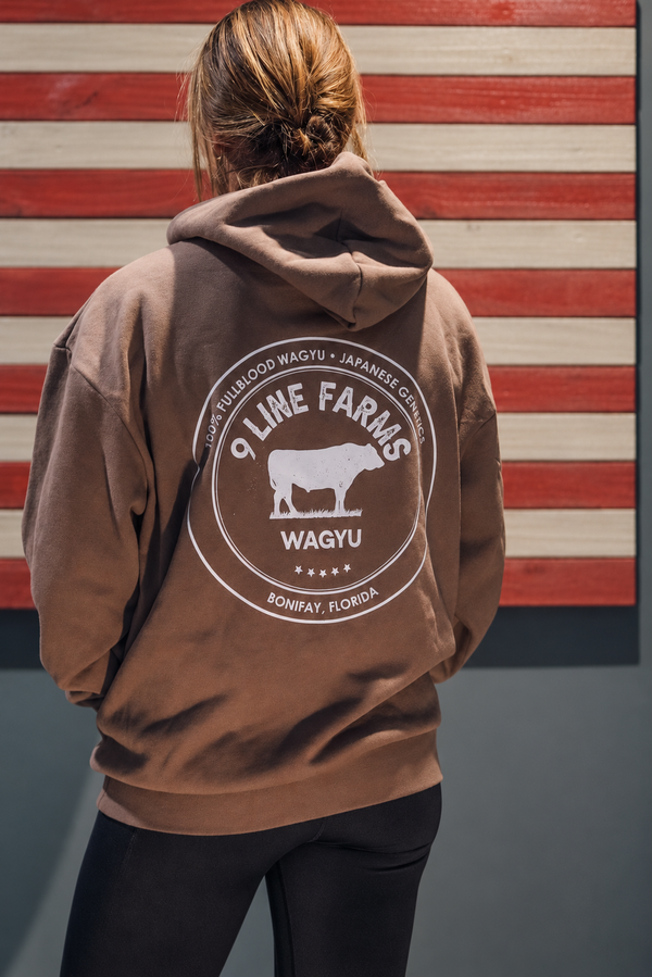 A person viewed from behind wearing a brown hoodie with a large circular farm graphic on the back that reads '9 Line Farms Wagyu Wagyu Farm Wagyu' and 'Wagyu, Florida.' The background shows a red, white, and gray striped wall.