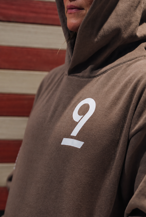 A close-up of a person wearing a brown hoodie with a large white number 9 printed on the chest, set against a background of red and beige horizontal wooden planks.
