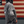 A person wearing a dark gray hooded sweatshirt stands in profile in front of a rustic American flag wall. The hoodie has a large white circular graphic on the back and a white American flag patch on the sleeve.