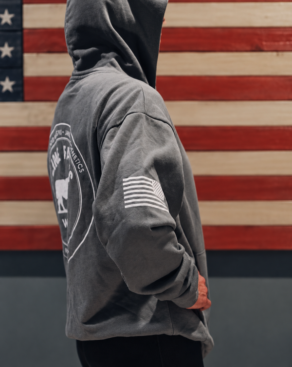 A person wearing a dark gray hooded sweatshirt stands in profile in front of a rustic American flag wall. The hoodie has a large white circular graphic on the back and a white American flag patch on the sleeve.