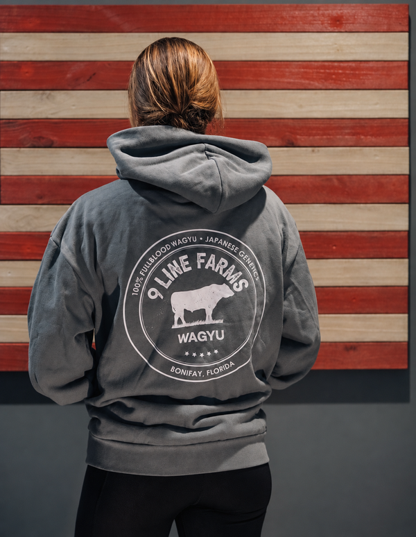 A person shown from behind wearing a dark gray hoodie with a large white circular graphic on the back that reads '9 Line Farms Wagyu' and 'Sonita, Florida,' standing in front of a rustic red-and-beige wood plank wall.