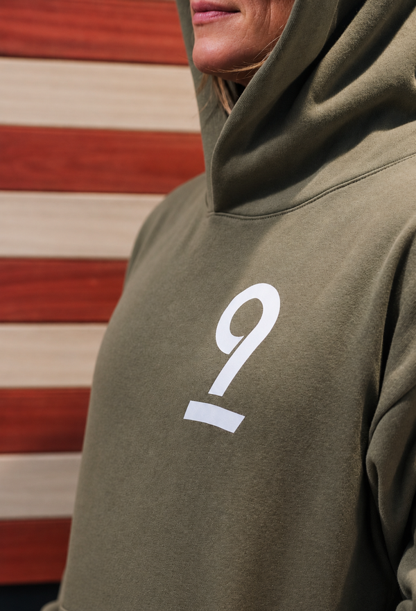 Close-up of a person wearing an olive green hoodie with a large white number 9 printed on the chest, set against a red and white striped wooden background.