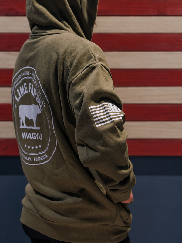 A person wearing an olive green hoodie, shown from the back and side. The hoodie features a large white circular '9 Line Farm Wagyu' graphic on the back and a white American flag patch on the sleeve.
